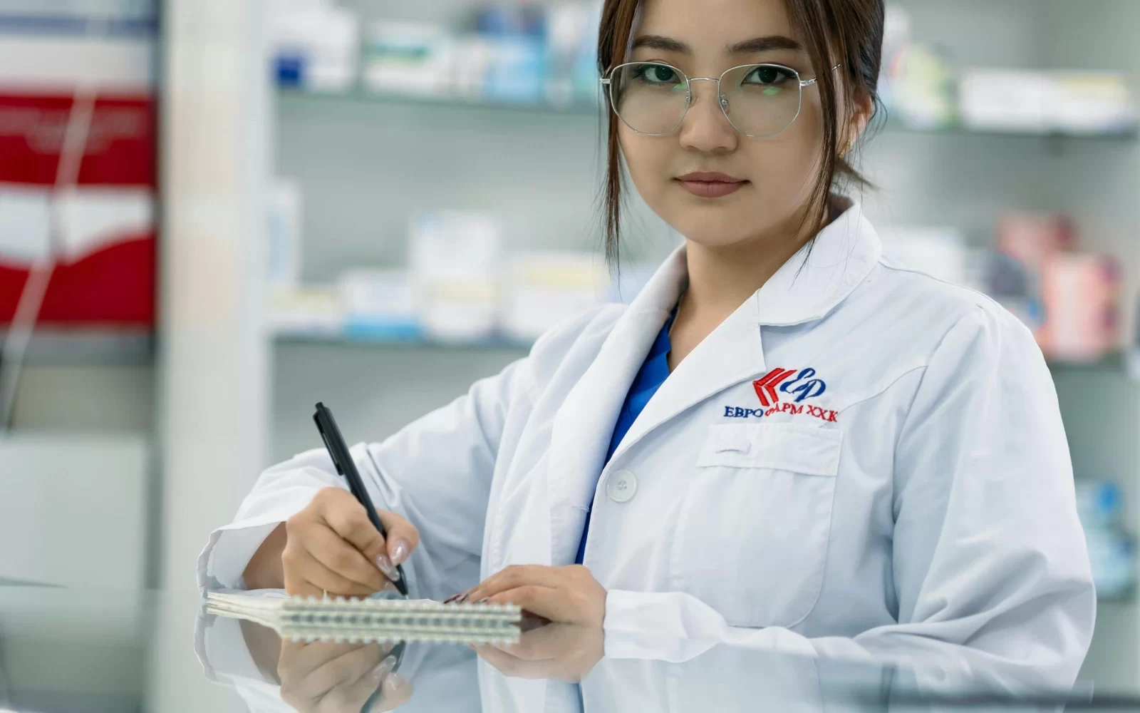Mujer con bata de laboratorio y gafas, tomando notas en un cuaderno en una farmacia, rodeada de productos farmacéuticos en estanterías.