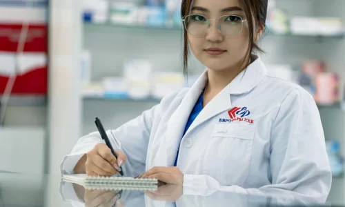 Mujer con bata de laboratorio y gafas, tomando notas en un cuaderno en una farmacia, rodeada de productos farmacéuticos en estanterías.