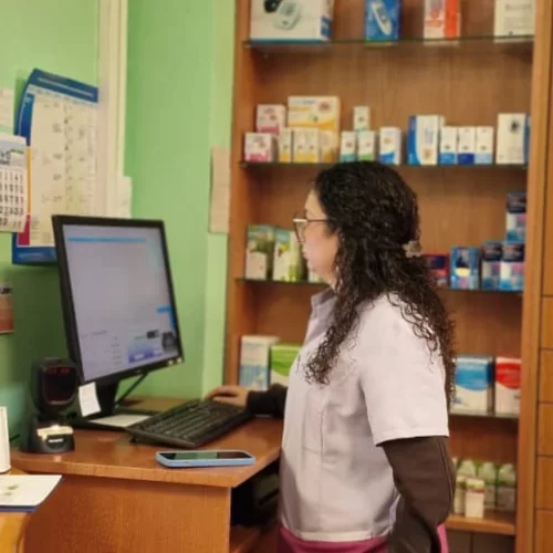 Farmacéutica trabajando en una computadora en una farmacia, rodeada de estantes con productos farmacéuticos y material de oficina.