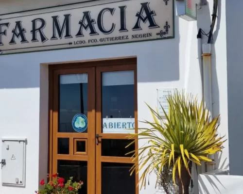 Fachada de una farmacia en Negrín, con un letrero que dice "FARMACIA" y una planta decorativa en la entrada. La puerta de madera está abierta, indicando que el establecimiento está disponible para atención al público.